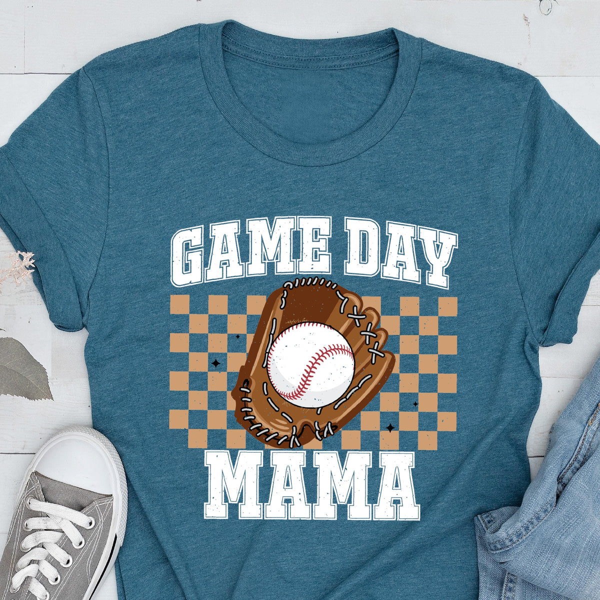 May include: A teal blue short-sleeve t-shirt featuring a graphic print with the text "GAME DAY MAMA" in white block letters. The design includes a brown baseball glove holding a white baseball, set against a tan and brown checkered background.