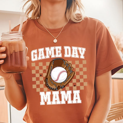 May include: A rust-colored short-sleeve t-shirt featuring a graphic print with the text GAME DAY MAMA in white block letters. The design includes a brown baseball glove holding a white baseball, centered over a tan and brown checkered pattern.