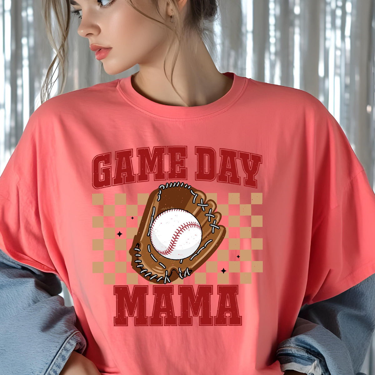 May include: A coral-colored t-shirt featuring a graphic print of a brown baseball glove holding a white baseball. The design includes a tan checkered background and the red text "GAME DAY MAMA" in a bold, athletic-style block font.