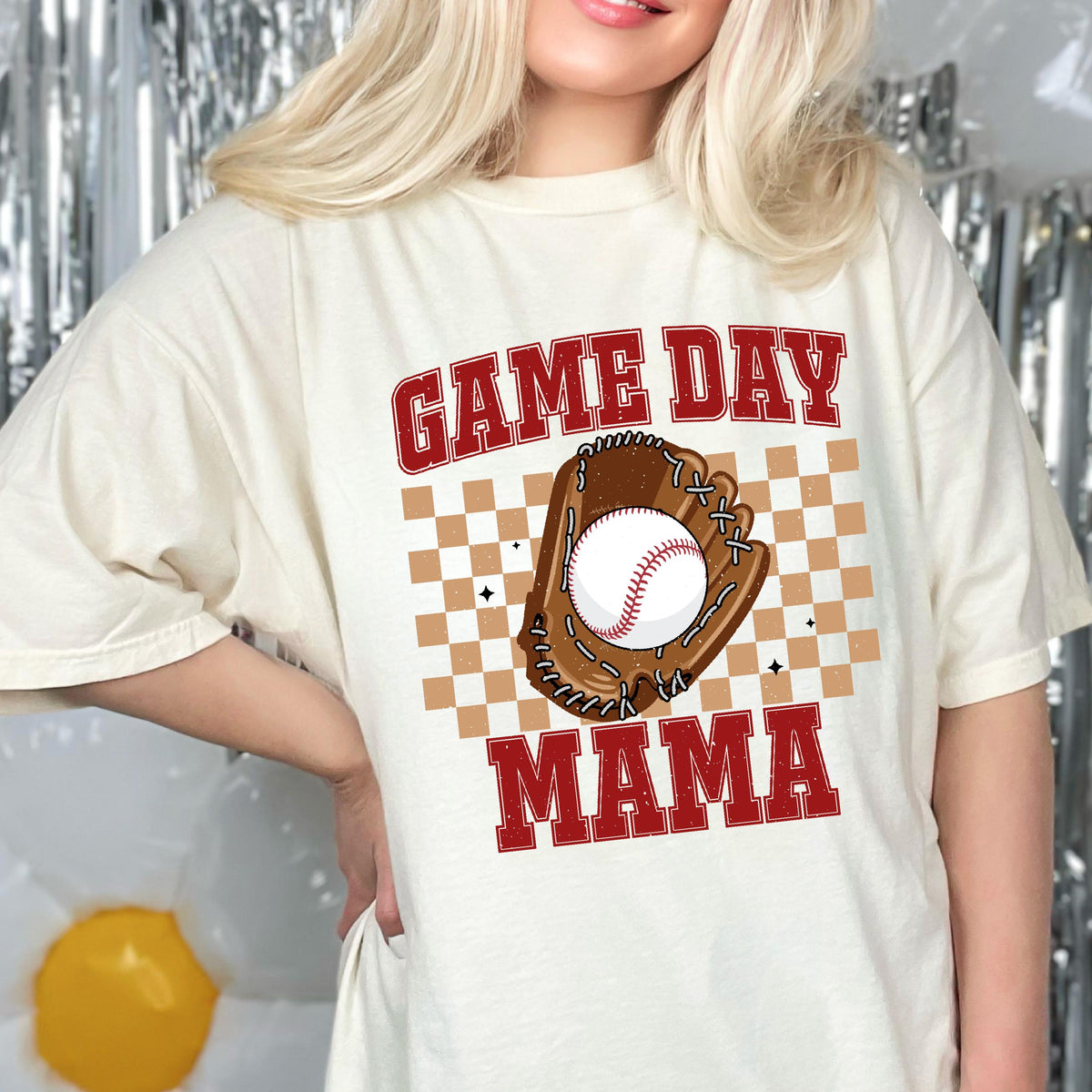 May include: An off-white short-sleeve t-shirt featuring a graphic print with the red text GAME DAY MAMA. The design includes a brown baseball glove holding a white baseball, centered over a tan and white checkered square pattern with small stars.