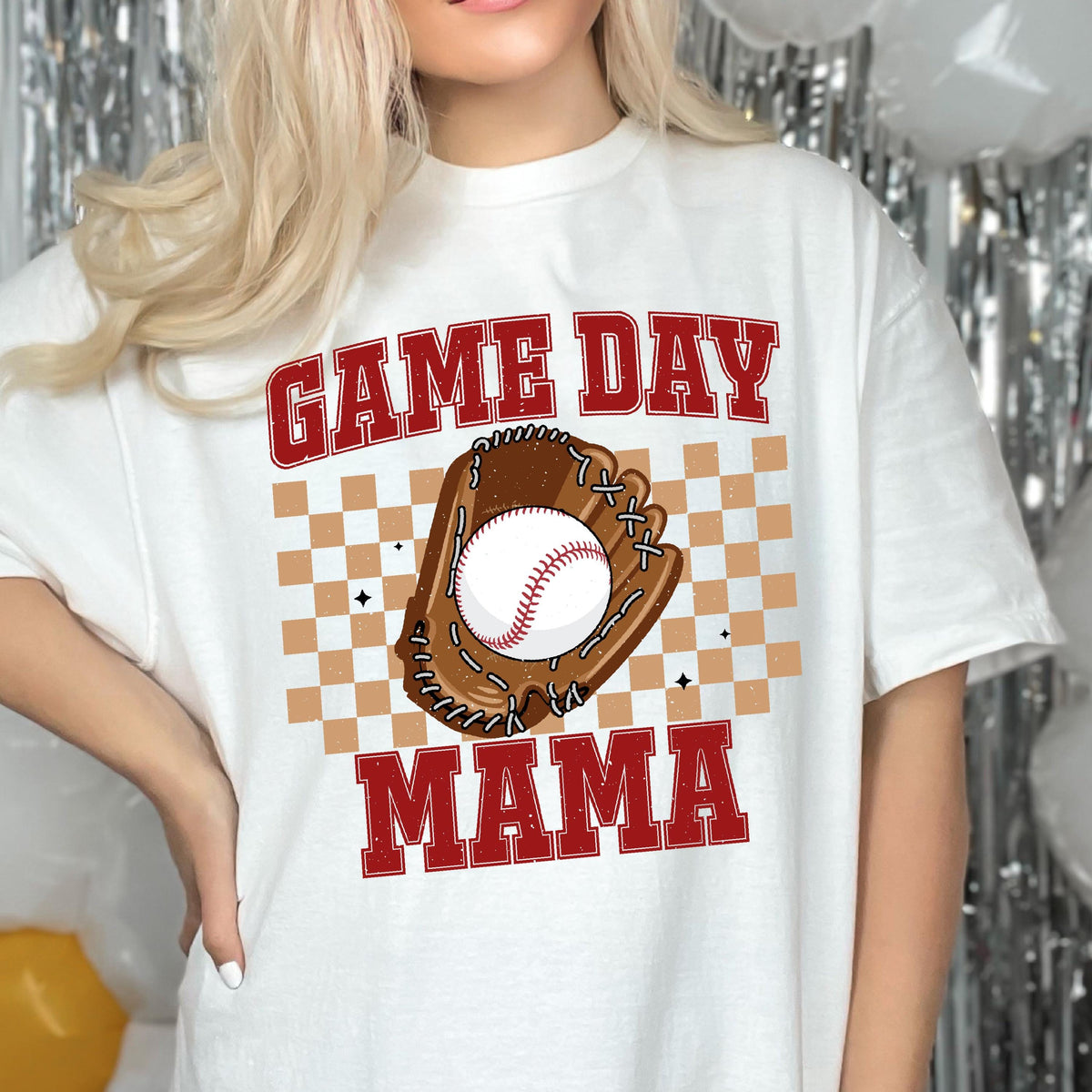 May include: A white t-shirt featuring a graphic print with the red text GAME DAY MAMA. The design includes a brown baseball glove holding a white baseball with red stitching, centered over a tan and white checkered square pattern background.