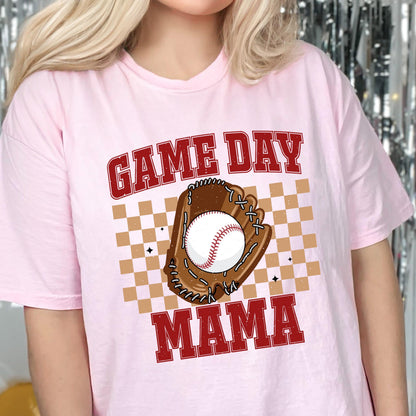 May include: A light pink t-shirt featuring a graphic print with the text GAME DAY MAMA in red block letters. The design includes a brown baseball glove holding a white baseball with red stitching, centered over a tan and white checkered square pattern.