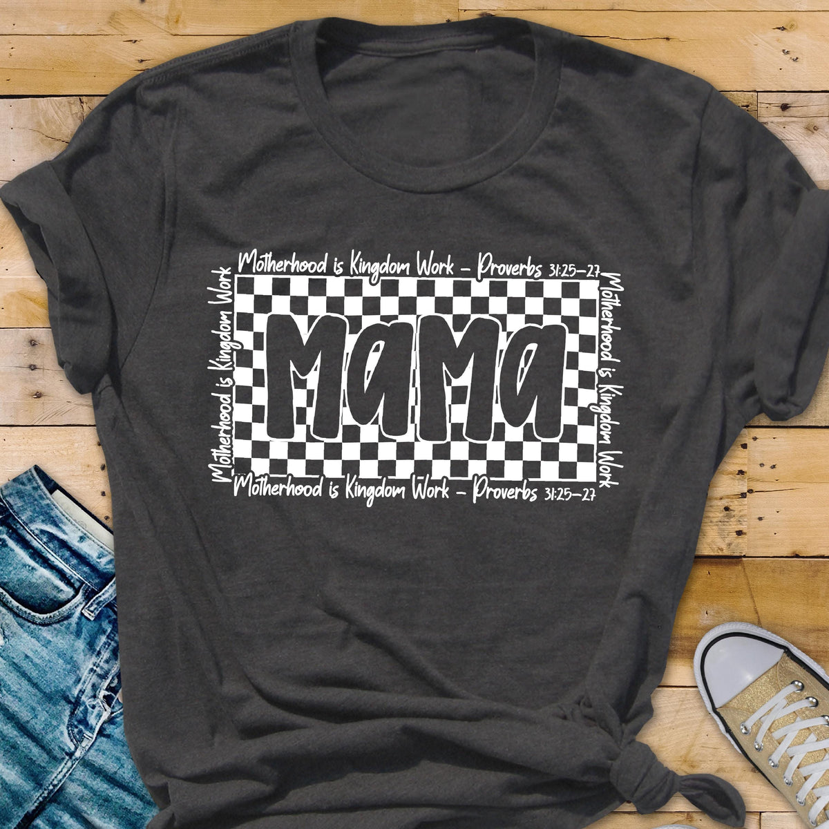 May include: A dark gray t-shirt with a white checkered design and the word "Mama" in bold white letters. The text "Motherhood is Kingdom Work - Proverbs 3:25-27" is repeated on the top and bottom of the checkered design.