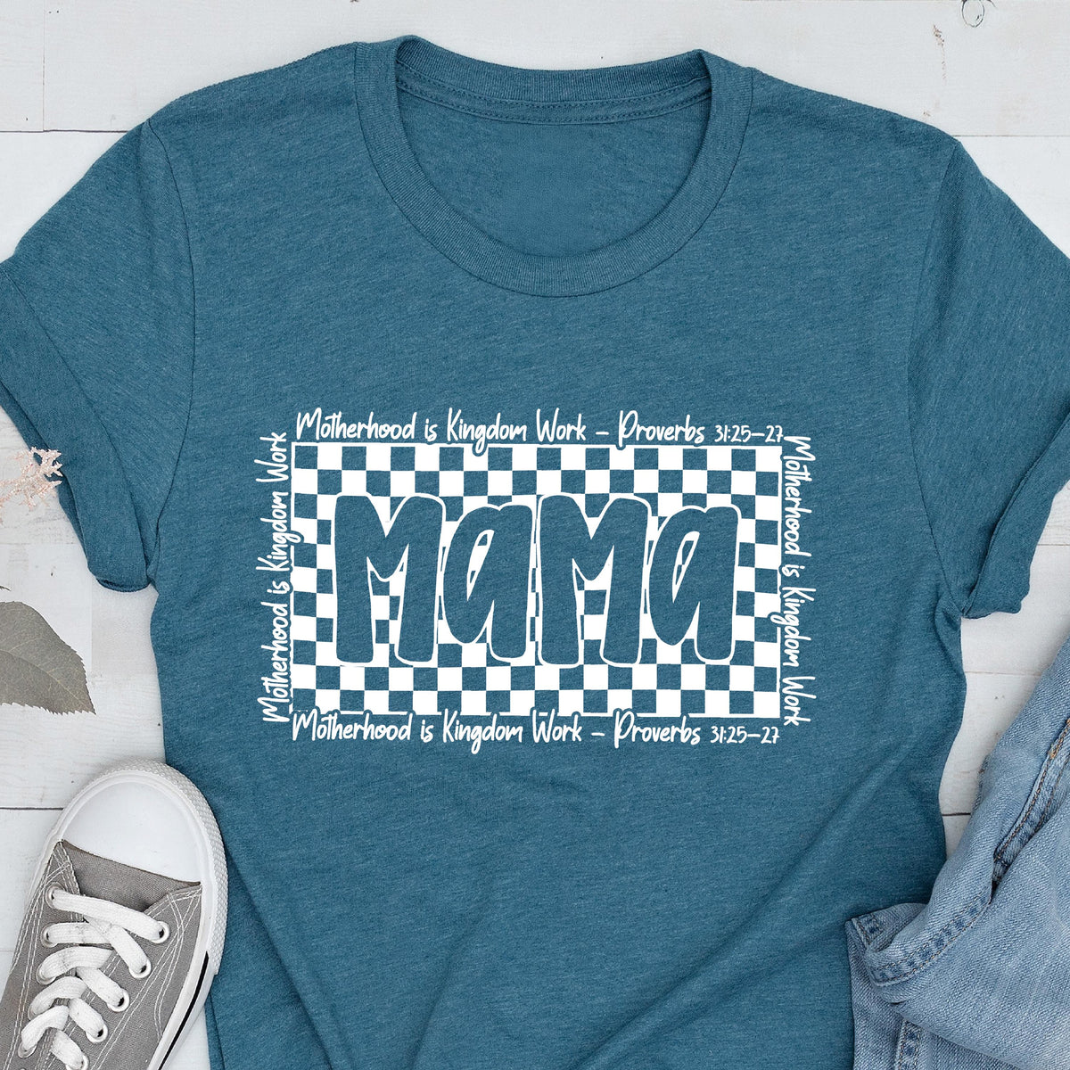 May include: Teal blue t-shirt with a white checkered background and the word "mama" in white outlined letters. The text "Motherhood is Kingdom Work - Proverbs 3:25-27" is repeated on the top and bottom of the checkered background.