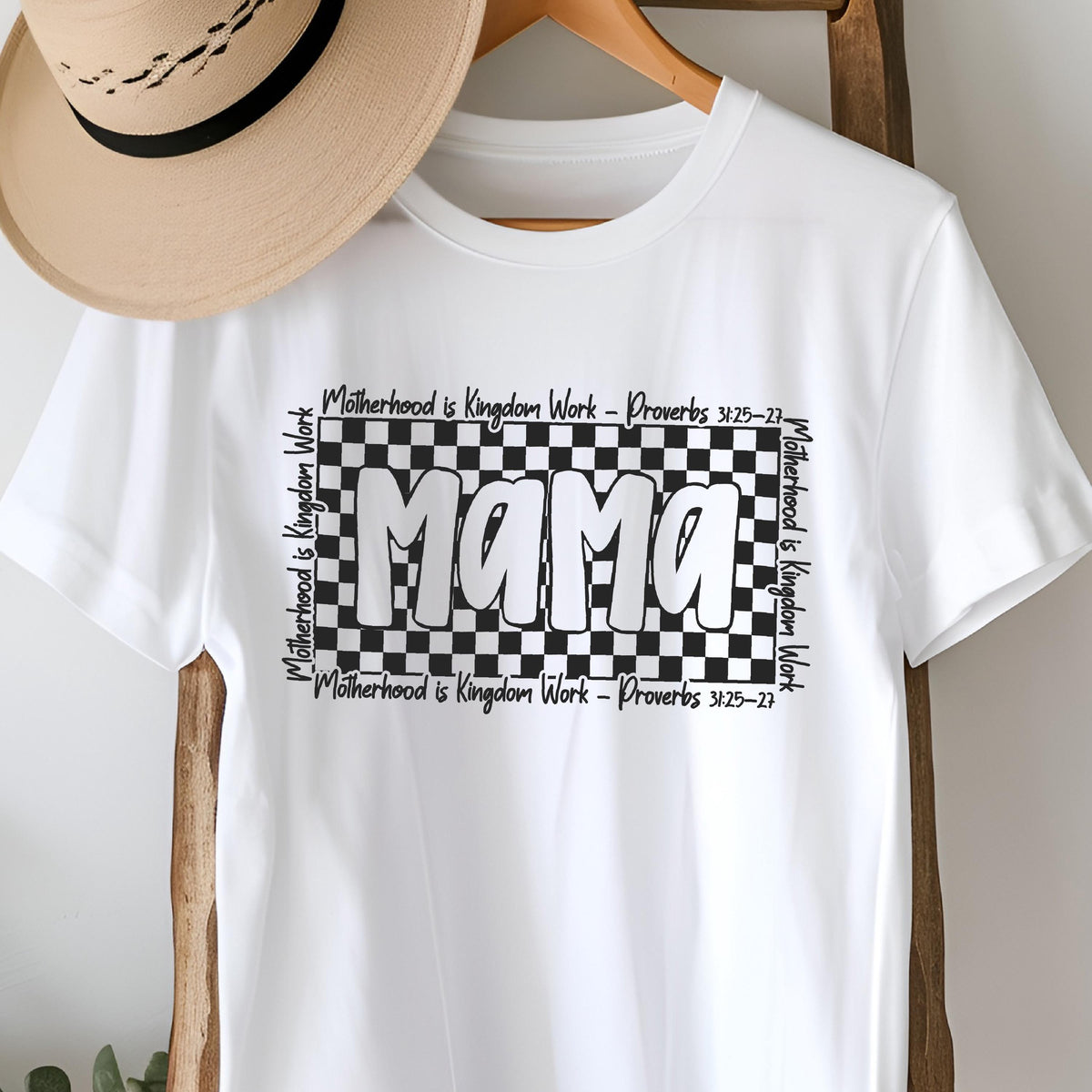 May include: White t-shirt with a black and white checkered design and the word "Mama" in bold, black letters. The text "Motherhood is Kingdom Work - Proverbs 3:25-27" is printed on the top and bottom of the checkered design.