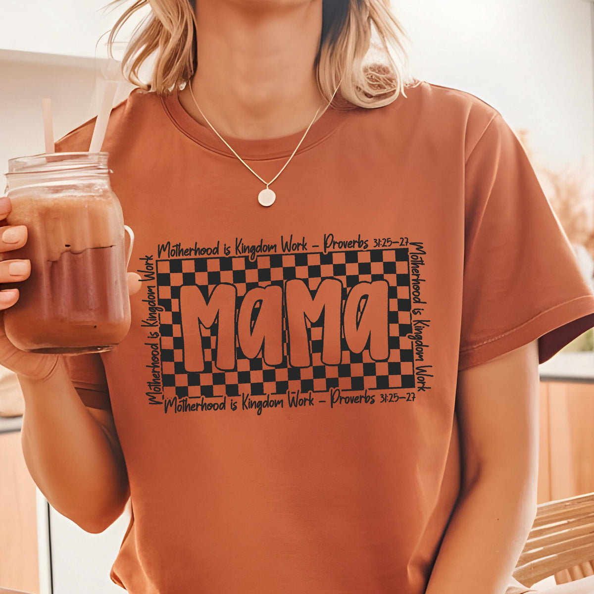 May include: A brown t-shirt with a black and white checkered design and the word "Mama" in black text. The text "Motherhood is Kingdom Work - Proverbs 3:25-27" is repeated on the top and bottom of the design.