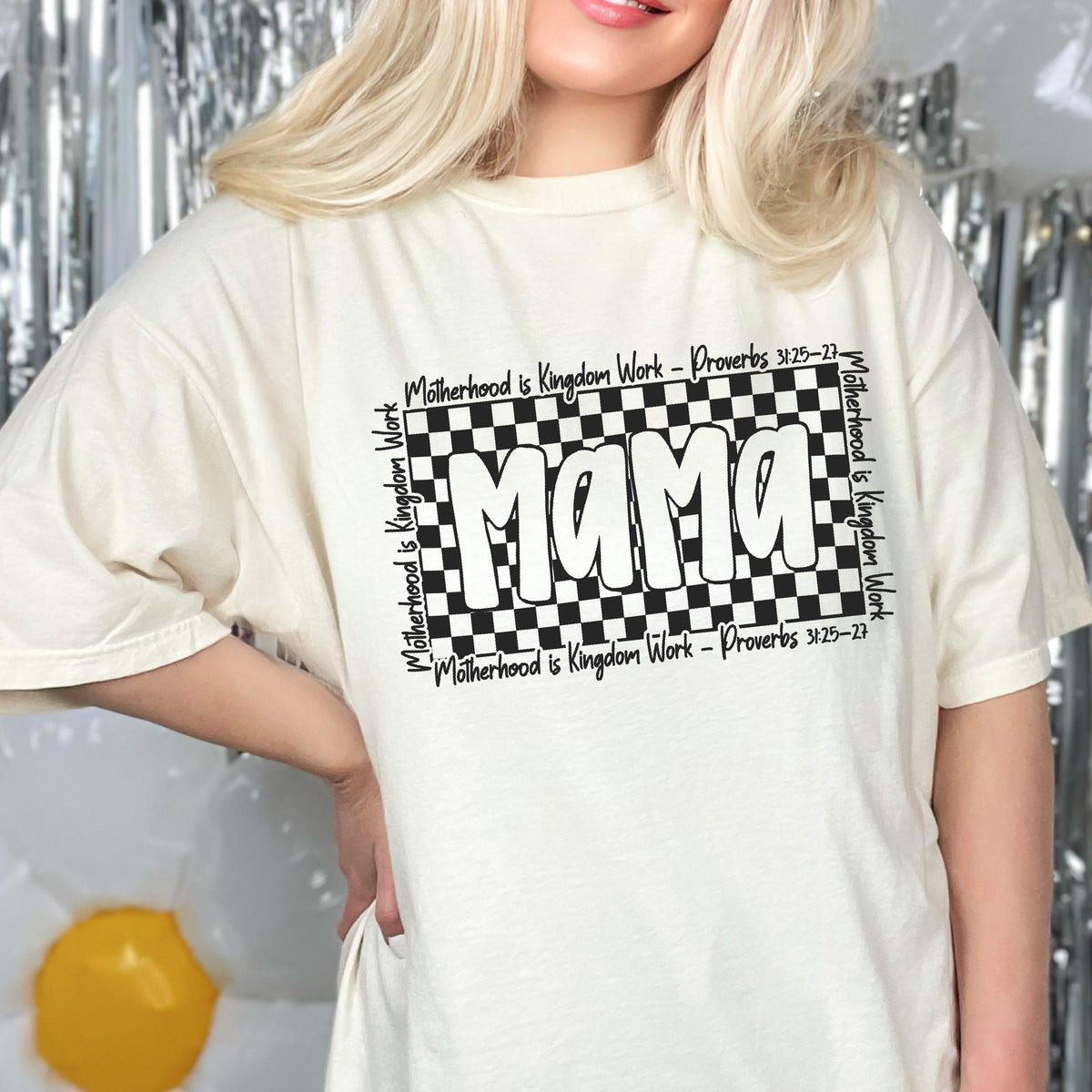May include: A white t-shirt with a black and white checkered design featuring the word "Mama" in bold white letters. The text "Motherhood is Kingdom Work - Proverbs 3:25-27" is repeated around the checkered design.