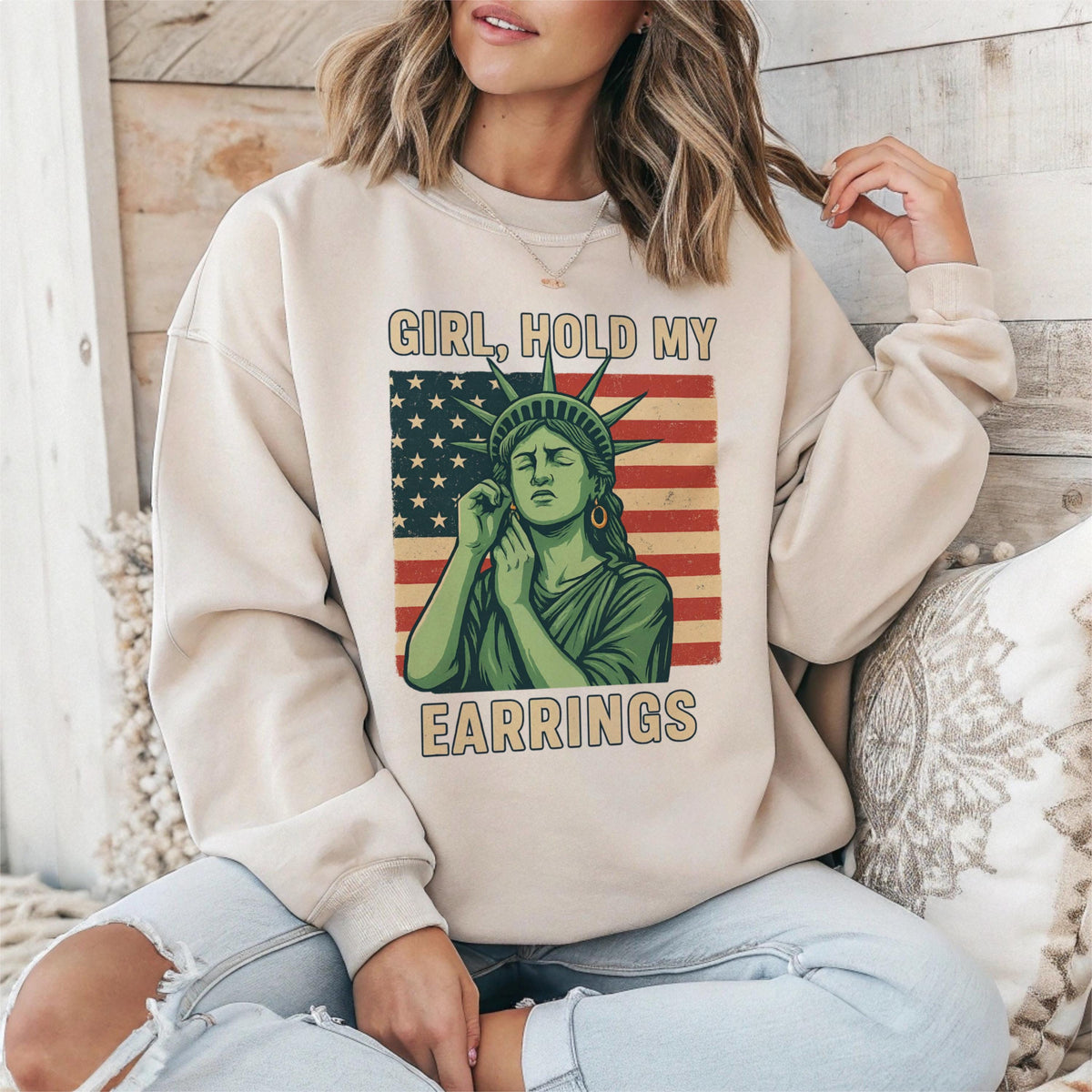 May include: A beige sweatshirt with a graphic print of the Statue of Liberty holding her ear and the text "Girl, Hold My Earrings".