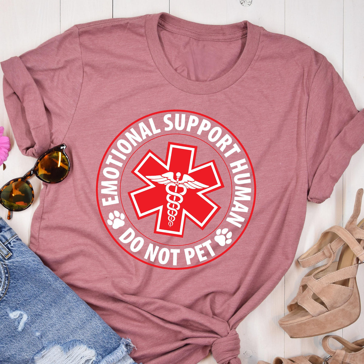 May include: A dusty rose t-shirt featuring a red and white circular design with the text "EMOTIONAL SUPPORT HUMAN DO NOT PET". The design includes a red medical symbol and paw prints. The shirt is folded, with sunglasses and sandals visible.