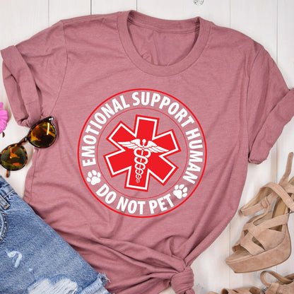 May include: A dusty rose t-shirt featuring a red and white circular design with the text "EMOTIONAL SUPPORT HUMAN DO NOT PET". The design includes a red medical symbol and paw prints. The shirt is folded, with sunglasses and sandals visible.