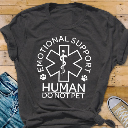 May include: A dark gray short-sleeved t-shirt featuring a white graphic of a Star of Life symbol with a snake and staff. The text reads EMOTIONAL SUPPORT HUMAN DO NOT PET, accompanied by two small paw print icons on either side of the central emblem.