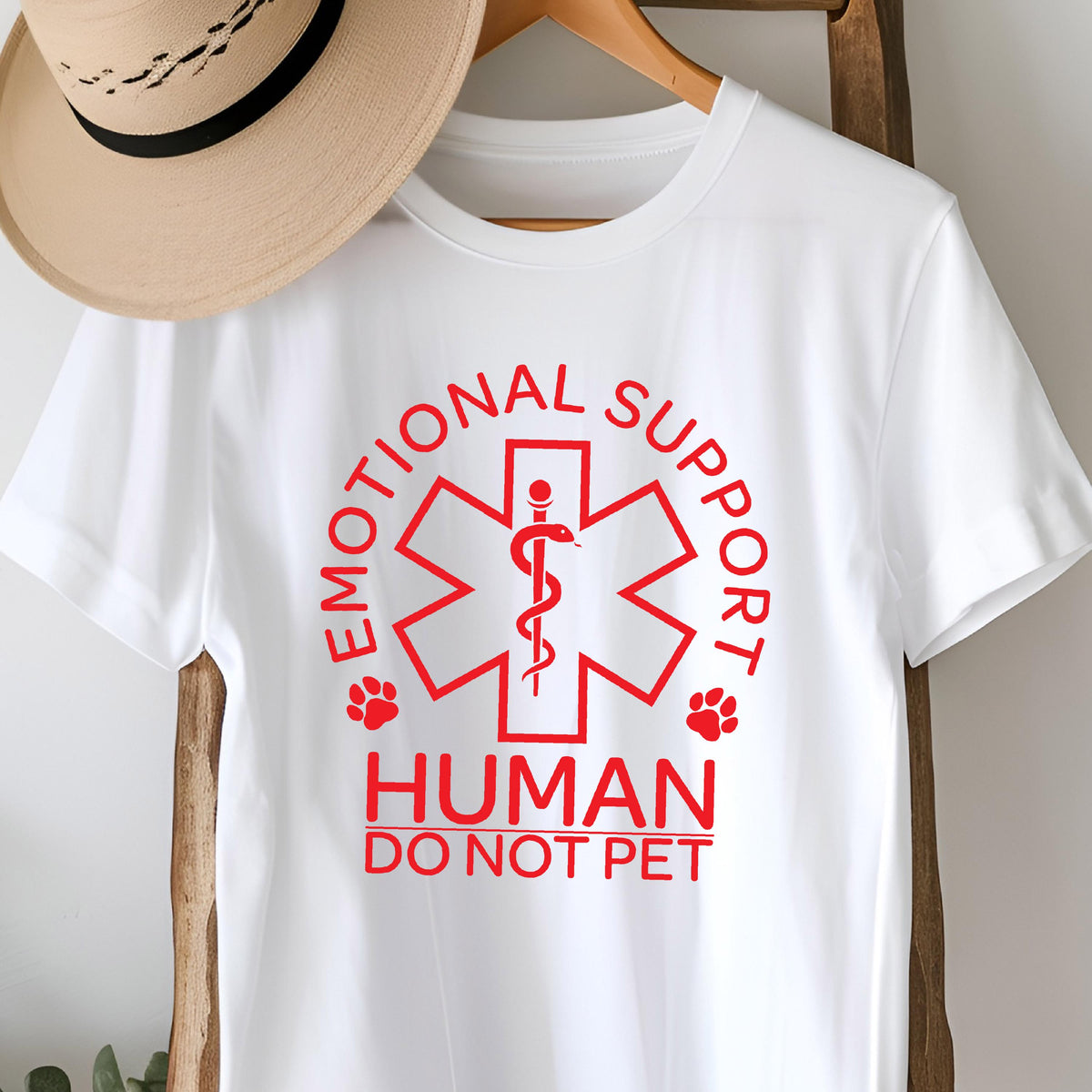 May include: A white short-sleeved t-shirt featuring a red graphic of a Star of Life symbol with a snake and staff. The text reads EMOTIONAL SUPPORT HUMAN DO NOT PET, accompanied by two small red paw prints on either side of the central design.