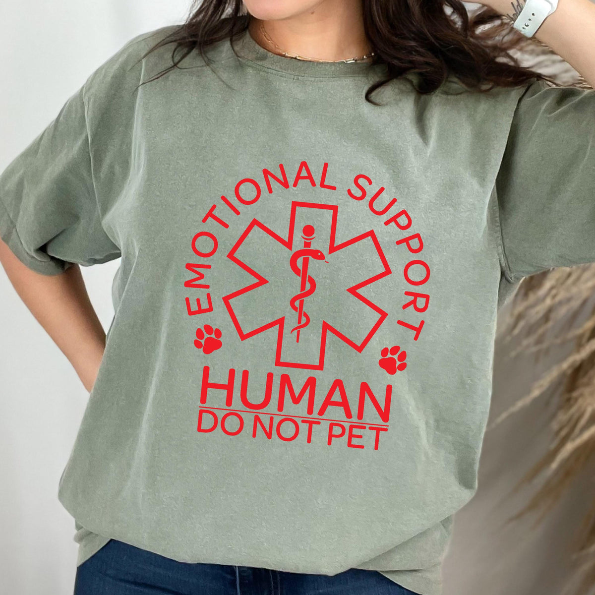 May include: A sage green short-sleeve t-shirt featuring a red graphic of a Star of Life symbol with a snake and staff. The text reads EMOTIONAL SUPPORT HUMAN DO NOT PET, accompanied by two small paw print icons on either side of the central emblem.