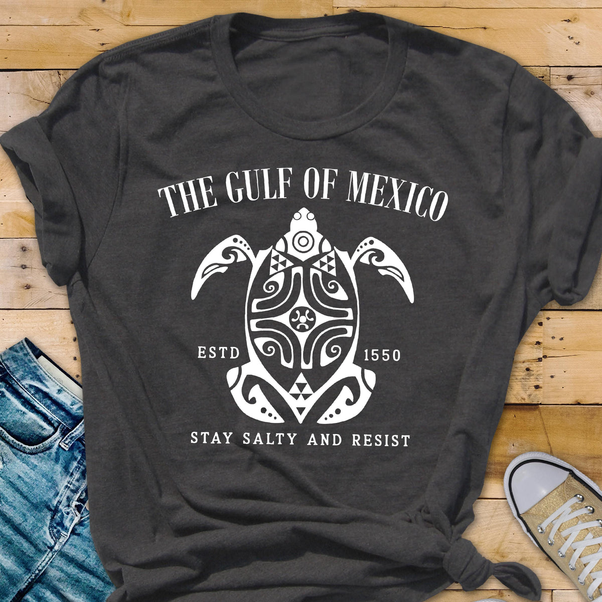 May include: A dark gray t-shirt with a white graphic design of a stylized turtle with the text "The Gulf of Mexico" above it and "Estd 1550" below it. The text "Stay Salty and Resist" is below the turtle.