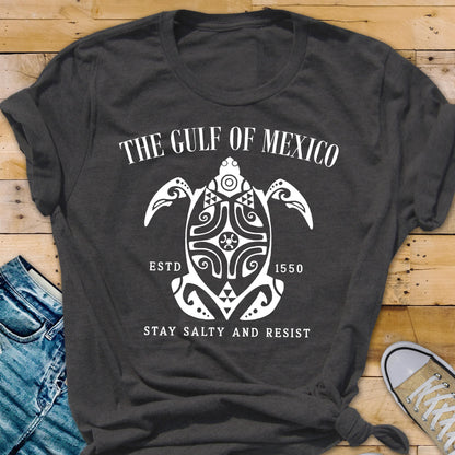 May include: A dark gray t-shirt with a white graphic design of a stylized turtle with the text "The Gulf of Mexico" above it and "Estd 1550" below it. The text "Stay Salty and Resist" is below the turtle.