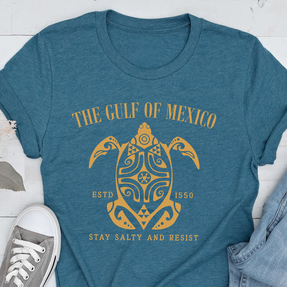 May include: A teal blue t-shirt with a gold graphic of a stylized turtle with the text "The Gulf of Mexico" above it and "Estd 1550" and "Stay Salty and Resist" below it.