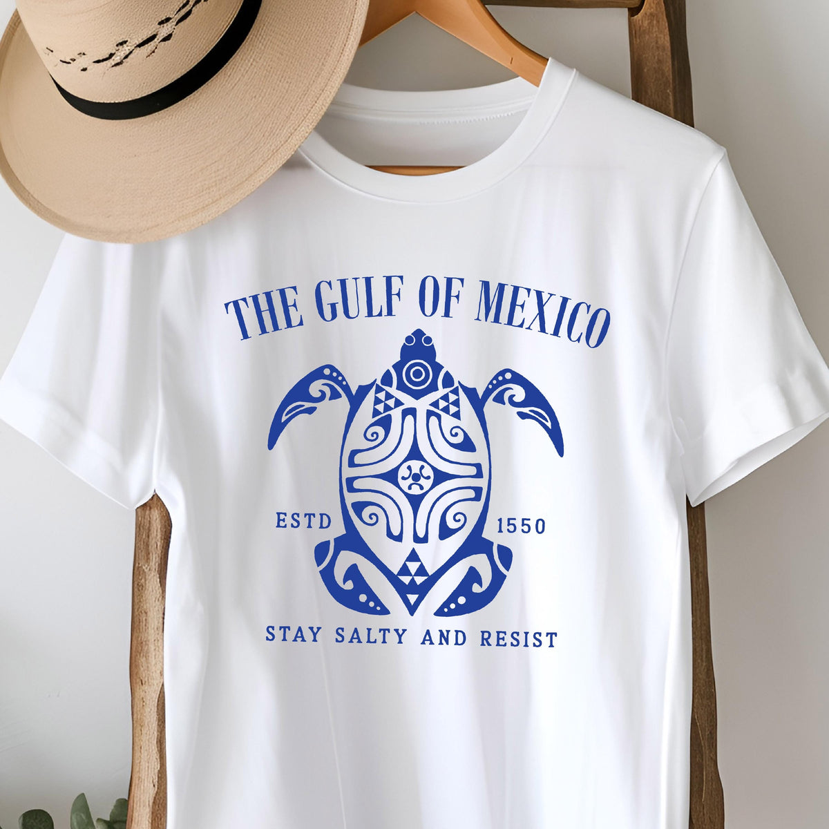 May include: White t-shirt with a blue graphic of a stylized turtle with the text "The Gulf of Mexico" above it, "Estd 1550" below it, and "Stay Salty and Resist" at the bottom.