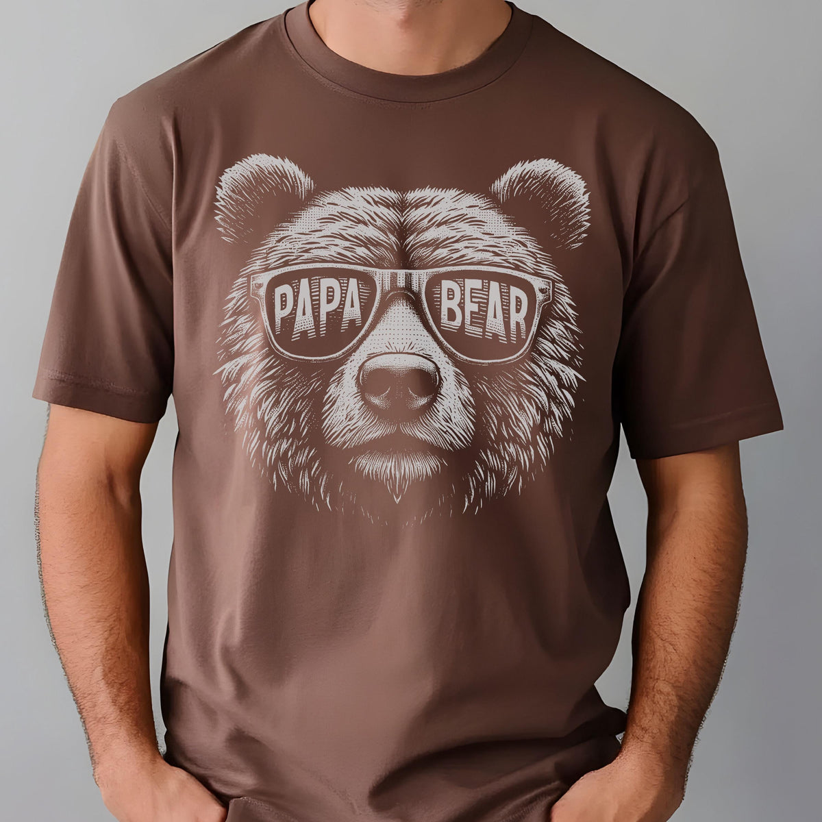 May include: Brown t-shirt featuring a detailed illustration of a bear's face wearing sunglasses. The sunglasses display the words "PAPA BEAR" in white. The shirt is a solid brown color.