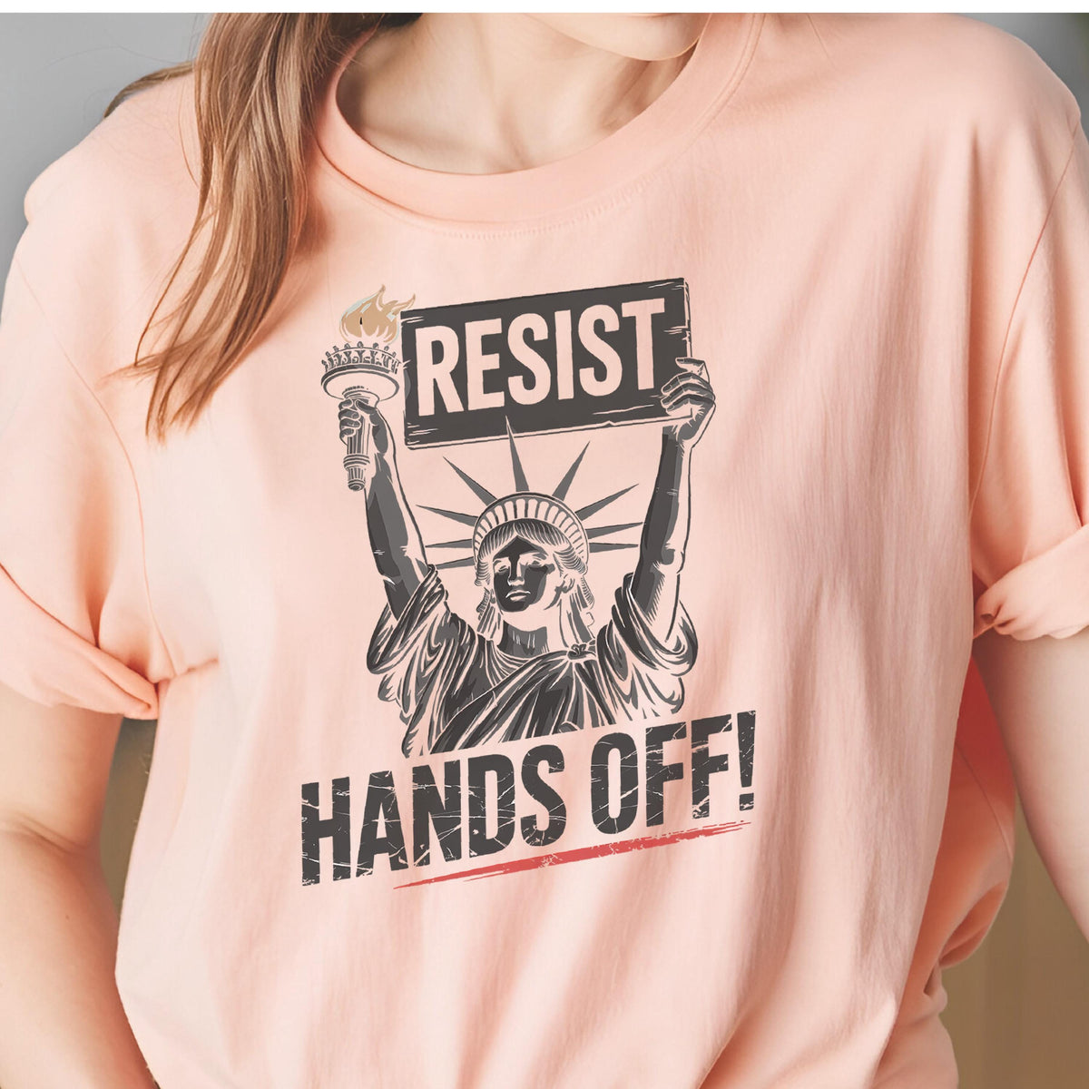 May include: A pink t-shirt with a graphic of the Statue of Liberty holding a sign that says "RESIST" and the text "HANDS OFF!" printed below.