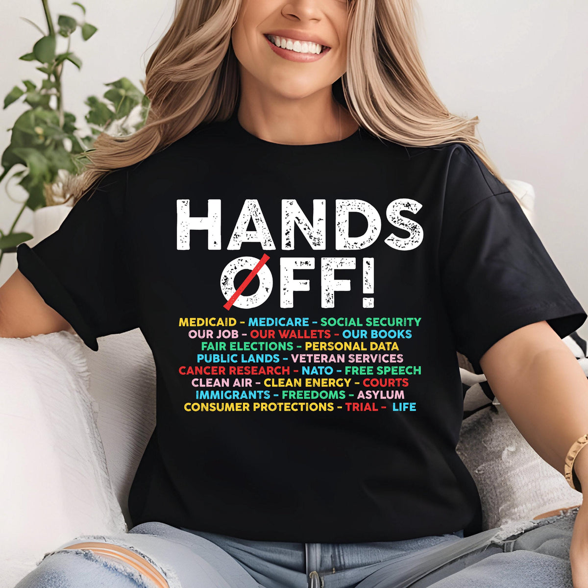 May include: Black t-shirt with the text "HANDS OFF!" in white. Below the text is a list of items including "Medicaid", "Medicare", "Social Security", "Our Job", "Our Wallets", "Our Books", "Fair Elections", "Personal Data", "Public Lands", "Veteran Services", "Cancer Research", "NATO", "Free Speech", "Clean Air", "Clean Energy", "Courts", "Immigrants", "Freedoms", "Asylum", "Consumer Protections", "Trial", and "Life".