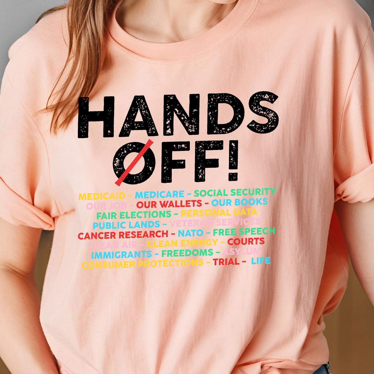 May include: A pink t-shirt with the text "HANDS OFF!" in black. Below the text is a list of items with a diagonal line through the word "our" in the phrase "our wallets". The list includes: Medicaid, Medicare, Social Security, Our Wallets, Our Books, Fair Elections, Personal Data, Public Lands, Veteran Services, Cancer Research, NATO, Free Speech, Clean Energy, Courts, Immigrants, Freedoms, Asylum, Consumer Protections, Trial, Life.