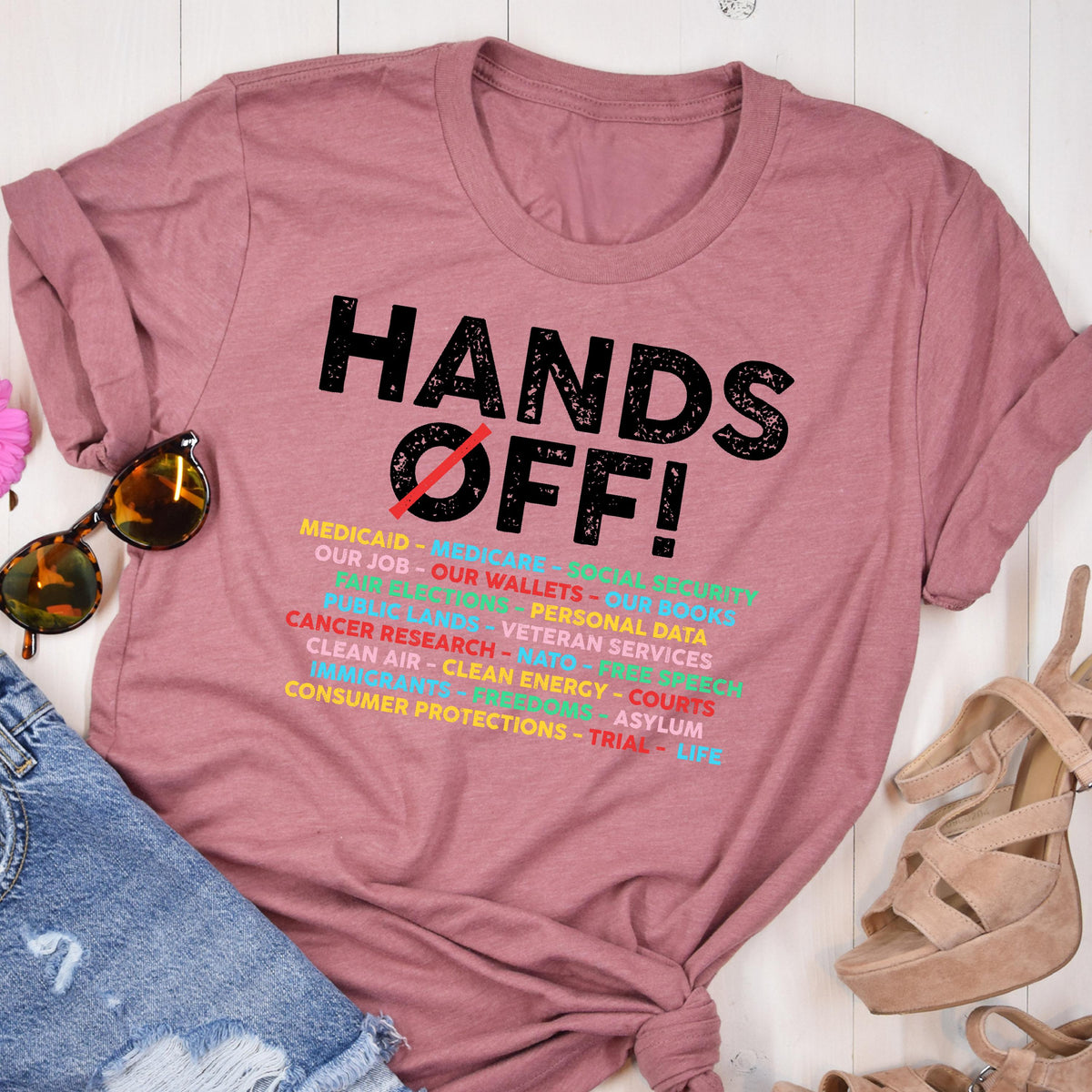 May include: A pink t-shirt with the text "HANDS OFF!" in black. The text is surrounded by a list of items in various colors, including "Medicaid", "Medicare", "Our Job", "Our Wallets", "Our Books", "Public Lands", "Veteran Services", "Personal Data", "Cancer Research", "NATO", "Free Speech", "Clean Air", "Clean Energy", "Courts", "Immigrants", "Asylum", "Consumer Protections", "Trial", and "Life".