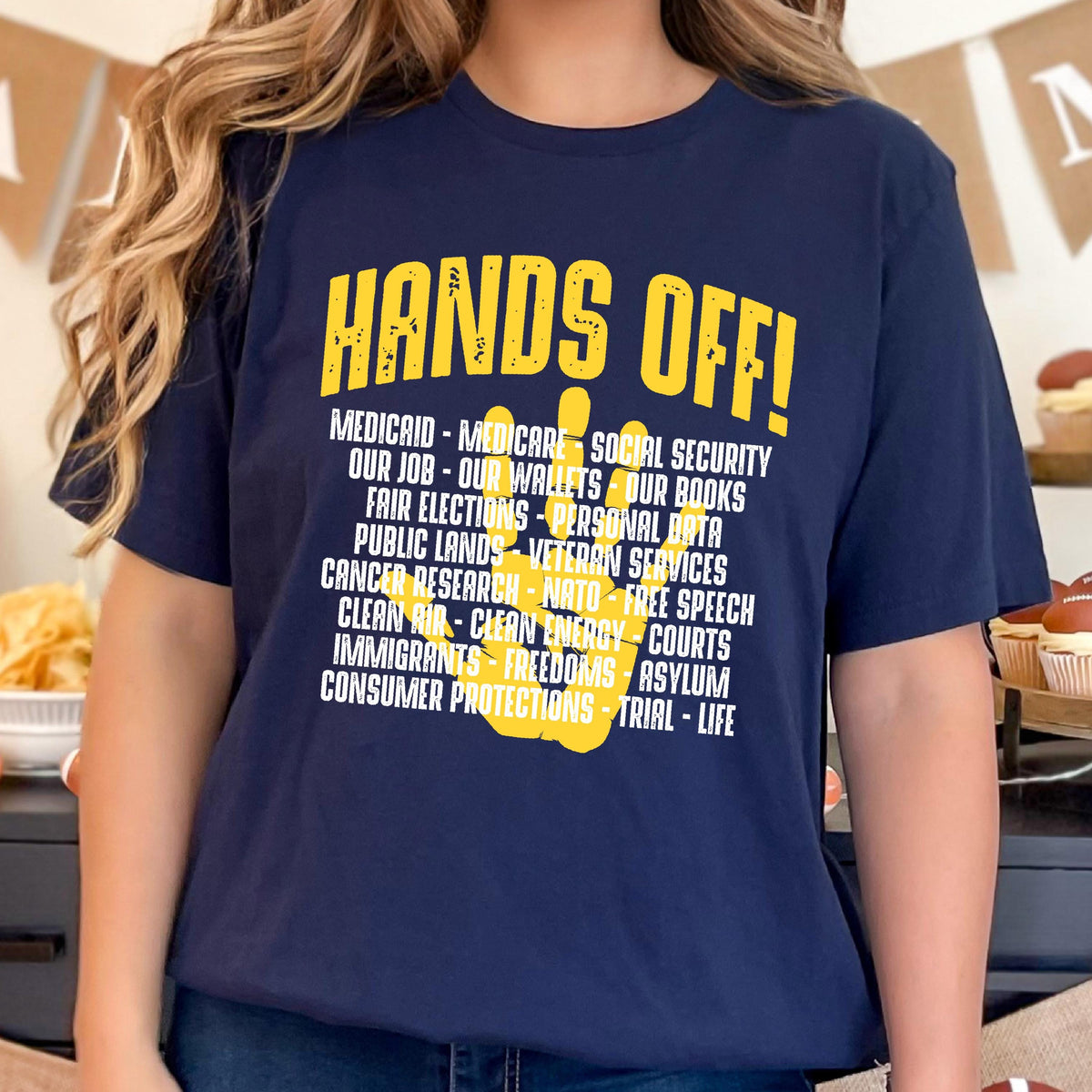 May include: A navy blue t-shirt with a yellow peace sign and the text "HANDS OFF!"  The text below the peace sign lists various social and political issues, including "Medicaid", "Medicare", "Social Security", "Our Job", "Our Wallets", "Our Books", "Fair Elections", "Personal Data", "Public Lands", "Veteran Services", "Cancer Research", "NATO", "Free Speech", "Clean Air", "Clean Energy", "Courts", "Immigrants", "Freedoms", "Asylum", "Consumer Protections", "Trial", and "Life".