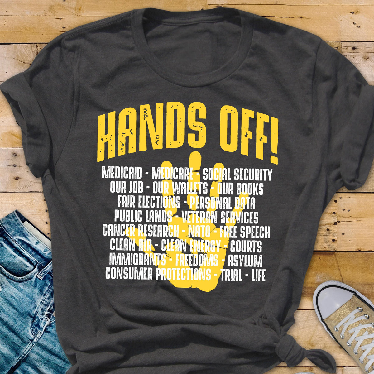 May include: A dark gray t-shirt with the text "HANDS OFF!" in yellow. The text is followed by a list of items including "Medicaid", "Medicare", "Social Security", "Our Job", "Our Wallets", "Our Books", "Fair Elections", "Personal Data", "Public Lands", "Veteran Services", "Cancer Research", "NATO", "Free Speech", "Clean Air", "Clean Energy", "Courts", "Immigrants", "Freedoms", "Asylum", "Consumer Protections", "Trial", and "Life".
