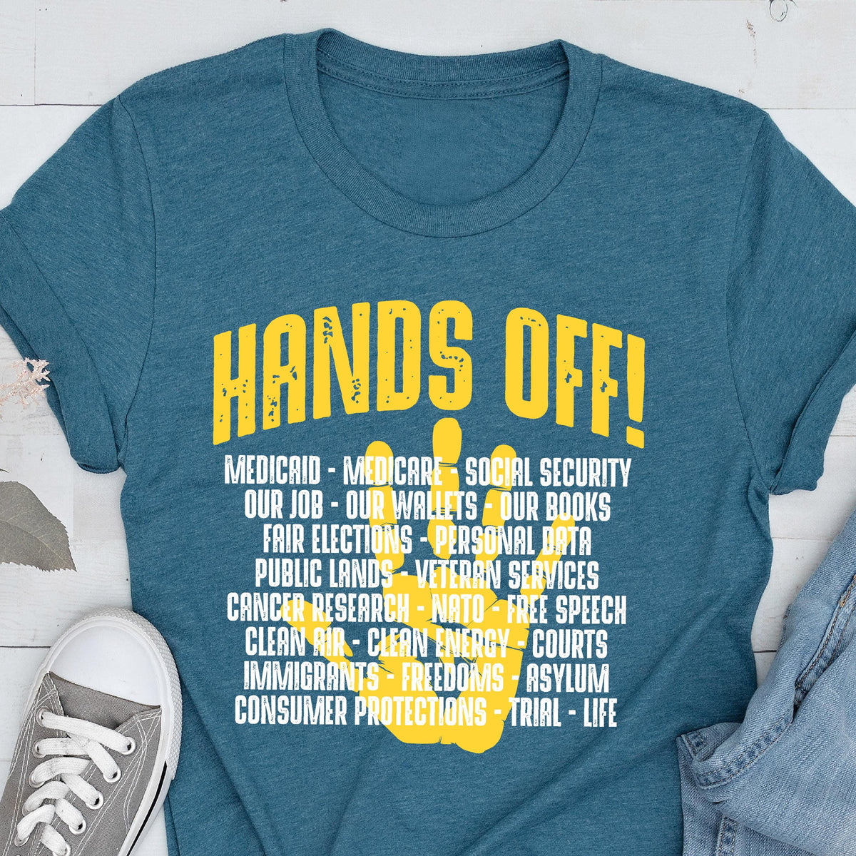 May include: A teal blue t-shirt with a yellow handprint graphic and the text "HANDS OFF!"  The handprint graphic has a list of items written on it, including "Medicaid", "Medicare", "Social Security", "Our Job", "Our Wallets", "Our Books", "Fair Elections", "Personal Data", "Public Lands", "Veteran Services", "Cancer Research", "NATO", "Free Speech", "Clean Air", "Clean Energy", "Courts", "Immigrants", "Freedoms", "Asylum", "Consumer Protections", "Trial", and "Life".