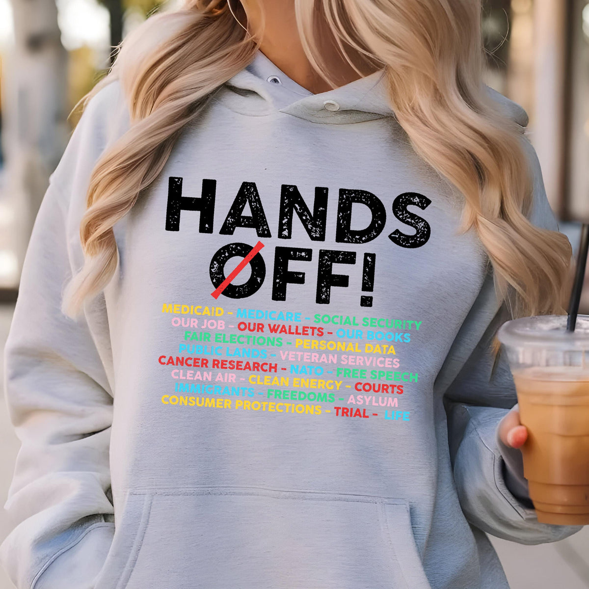 May include: A gray hoodie with the text "HANDS OFF!" in black. Below the text is a list of items including "Medicaid", "Medicare", "Social Security", "Our Job", "Our Wallets", "Our Books", "Fair Elections", "Personal Data", "Veteran Services", "Cancer Research", "NATO", "Free Speech", "Clean Air", "Clean Energy", "Courts", "Freedoms", "Asylum", "Consumer Protections", "Trial", and "Jury".