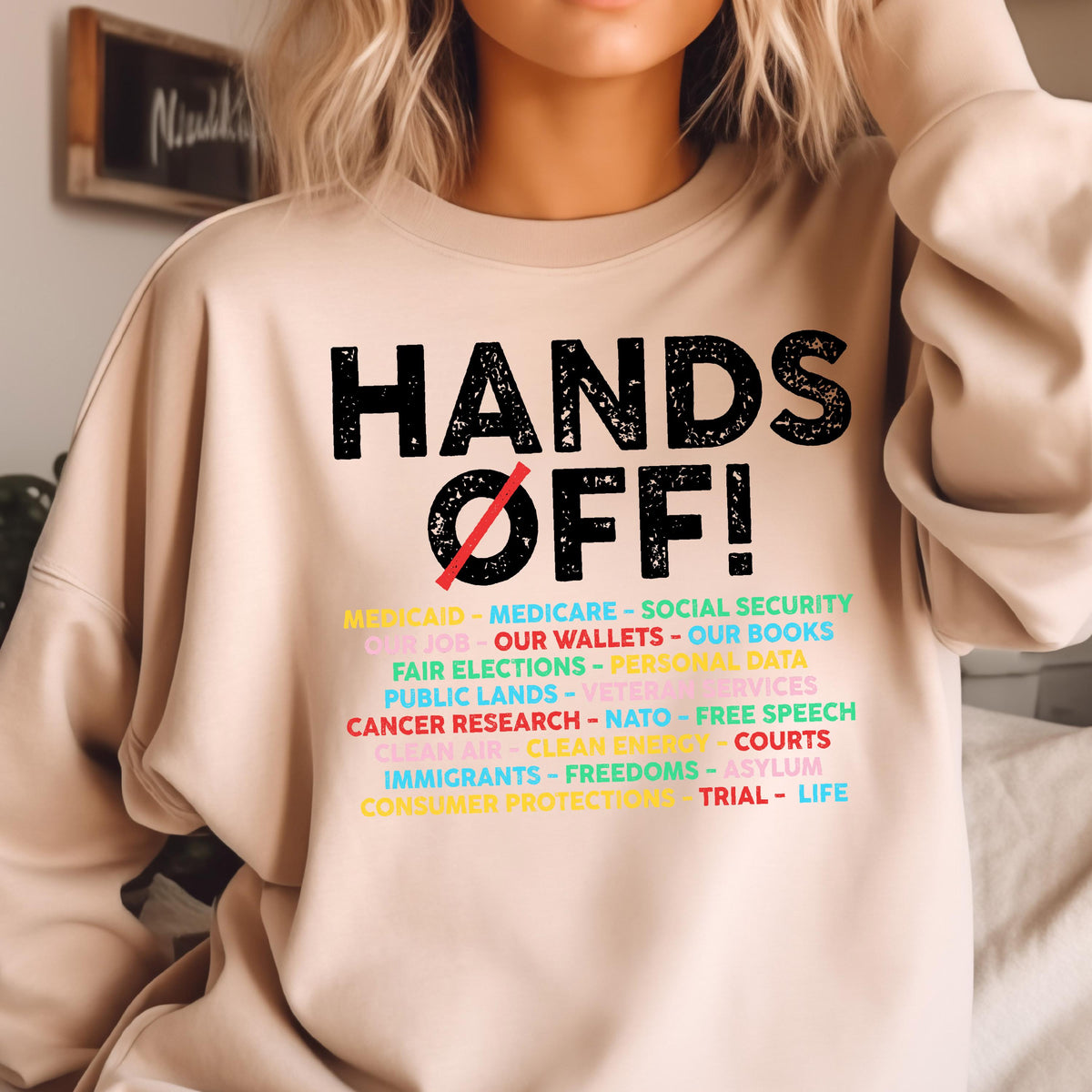 May include: A beige sweatshirt with the text "HANDS OFF!" in black. Below the text is a list of items in various colors, including "Medicaid", "Medicare", "Social Security", "Our Job", "Our Wallets", "Our Books", "Fair Elections", "Personal Data", "Public Lands", "Veterans Services", "Cancer Research", "NATO", "Free Speech", "Clean Air", "Clean Energy", "Courts", "Immigrants", "Freedoms", "Asylum", "Consumer Protections", "Trial", and "Life".