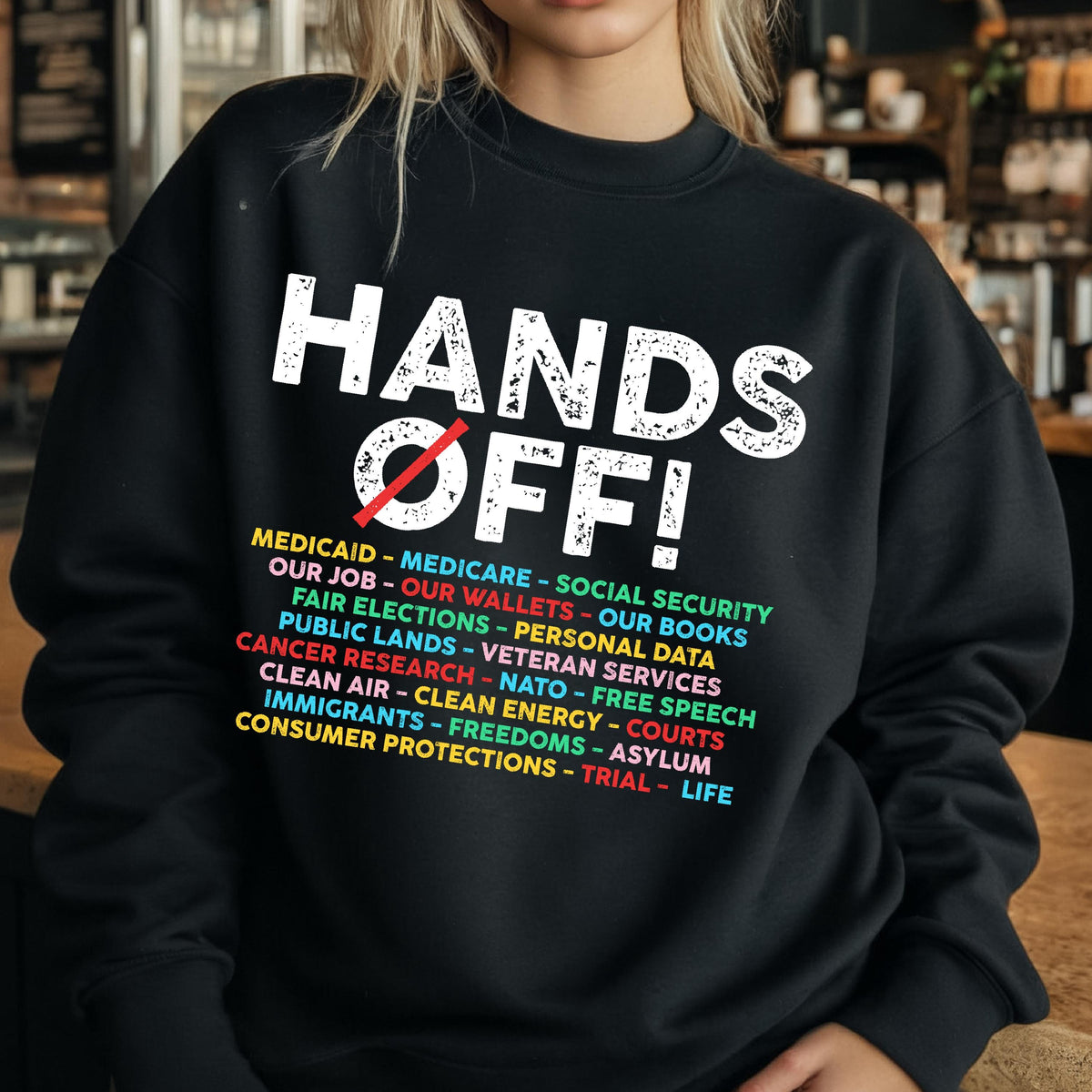 May include: Black sweatshirt with white text that reads "Hands Off!" and a list of items including "Medicaid", "Medicare", "Social Security", "Our Job", "Our Wallets", "Our Books", "Fair Elections", "Personal Data", "Public Lands", "Veteran Services", "Cancer Research", "NATO", "Free Speech", "Clean Air", "Clean Energy", "Courts", "Immigrants", "Freedoms", "Asylum", "Consumer Protections", "Trial", and "Life".