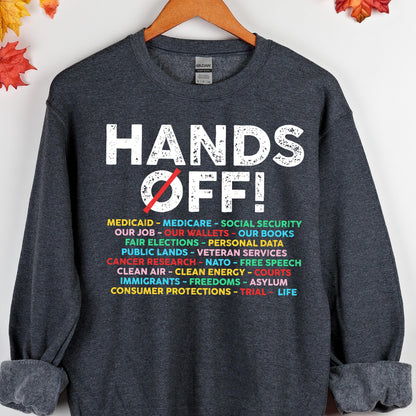 May include: A dark gray sweatshirt with the text "HANDS OFF!" in white. Below the text is a list of items including "Medicaid", "Medicare", "Social Security", "Our Job", "Our Wallets", "Our Books", "Fair Elections", "Personal Data", "Public Lands", "Veteran Services", "Cancer Research", "NATO", "Free Speech", "Clean Air", "Clean Energy", "Courts", "Immigrants", "Freedoms", "Asylum", "Consumer Protections", "Trial", and "Life".
