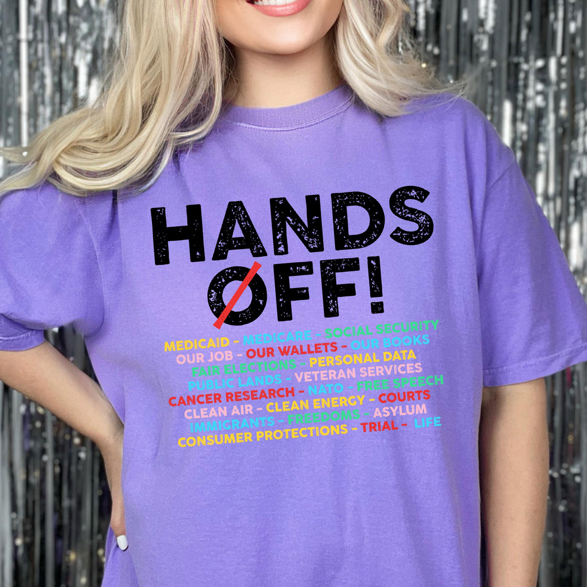 May include: A light purple short-sleeve t-shirt featuring the black text HANDS OFF! with a red diagonal line through the O. Below, a list of multi-colored words includes MEDICAID, MEDICARE, SOCIAL SECURITY, OUR JOB, OUR WALLETS, OUR BOOKS, and more.