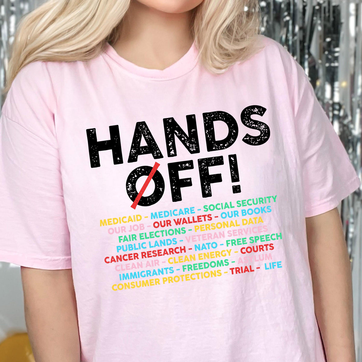 May include: A light pink t-shirt featuring the black text HANDS OFF! with a red slash through the O. Below the text is a list of various social and political topics printed in multiple colors including yellow, teal, red, and light pink.