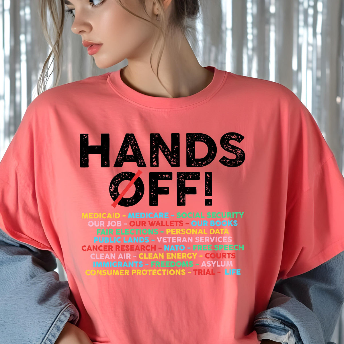 May include: A coral-colored t-shirt featuring the black text HANDS OFF! with a red slash through the O. Below, a list of multi-colored words includes MEDICAID, MEDICARE, SOCIAL SECURITY, OUR JOB, OUR WALLETS, OUR BOOKS, FAIR ELECTIONS, and more.