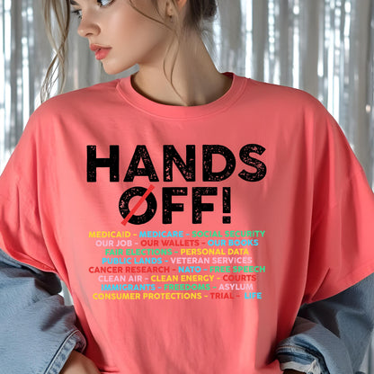 May include: A coral-colored t-shirt featuring the black text HANDS OFF! with a red slash through the O. Below, a list of multi-colored words includes MEDICAID, MEDICARE, SOCIAL SECURITY, OUR JOB, OUR WALLETS, OUR BOOKS, FAIR ELECTIONS, and more.