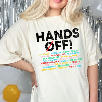 May include: A cream-colored short-sleeve t-shirt featuring the black text HANDS OFF! with a red slash through the O. Below, a list of terms including Medicaid, Medicare, Social Security, Our Job, Our Wallets, Our Books, Fair Elections, and more.