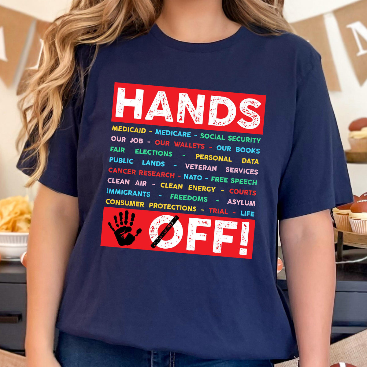 May include: A navy blue t-shirt with the text "HANDS OFF!" in red and white. The text is surrounded by a list of items in various colors, including "Medicaid", "Medicare", "Social Security", "Our Job", "Our Wallets", "Our Books", "Fair Elections", "Personal Data", "Public Lands", "Veteran Services", "Cancer Research", "NATO", "Free Speech", "Clean Air", "Clean Energy", "Courts", "Immigrants", "Freedoms", "Asylum", "Consumer Protections", "Trial", and "Life". A red hand with a diagonal line through it is ab