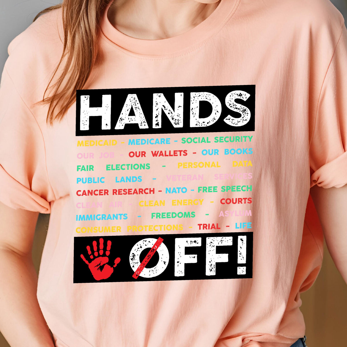 May include: Pink t-shirt with the text "HANDS OFF!" in black. The text is surrounded by a list of items in various colors, including "Medicare", "Social Security", "Our Wallets", "Our Books", "Fair Elections", "Public Lands", "Veteran Services", "Cancer Research", "NATO", "Free Speech", "Clean Air", "Clean Energy", "Courts", "Immigrants", "Freedoms", "Asylum", "Consumer Protections", "Trial", and "Life". A red handprint with a diagonal line through it is below the text.