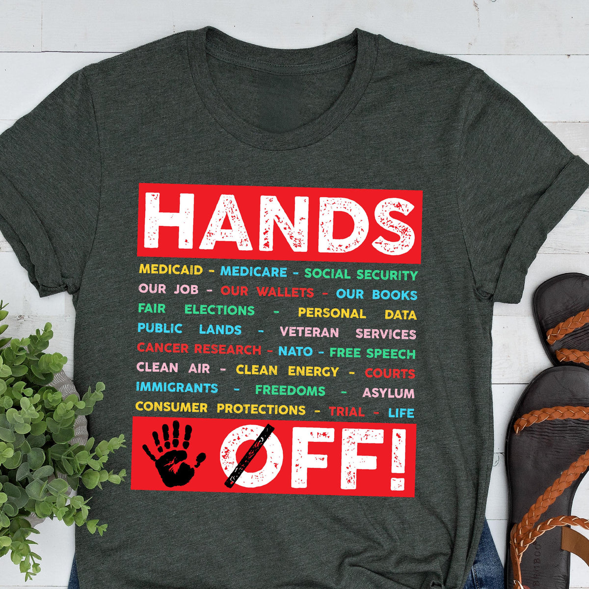 May include: A dark green t-shirt with a red graphic that says "Hands Off!" with a list of items underneath, including "Medicaid", "Medicare", "Social Security", "Our Job", "Our Wallets", "Our Books", "Fair Elections", "Personal Data", "Public Lands", "Veteran Services", "Cancer Research", "NATO", "Free Speech", "Clean Air", "Clean Energy", "Courts", "Immigrants", "Freedoms", "Asylum", "Consumer Protections", "Trial", and "Life".