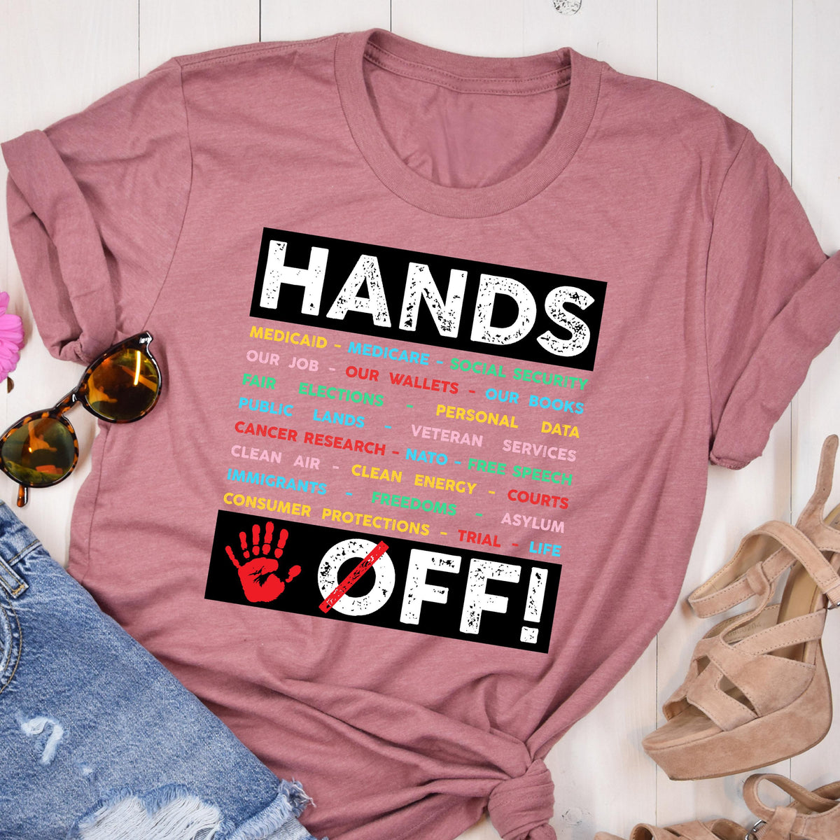 May include: A pink t-shirt with the text "HANDS OFF!" in black and white. The text is surrounded by a black rectangle with a list of words in different colors, including "Medicaid", "Medicare", "Our Job", "Our Wallets", "Our Books", "Personal Data", "Public Lands", "Veteran Services", "Cancer Research", "NATO", "Free Speech", "Clean Air", "Clean Energy", "Courts", "Immigrants", "Asylum", "Consumer Protections", "Trial", and "Life". A red hand with a diagonal line through it is below the text.