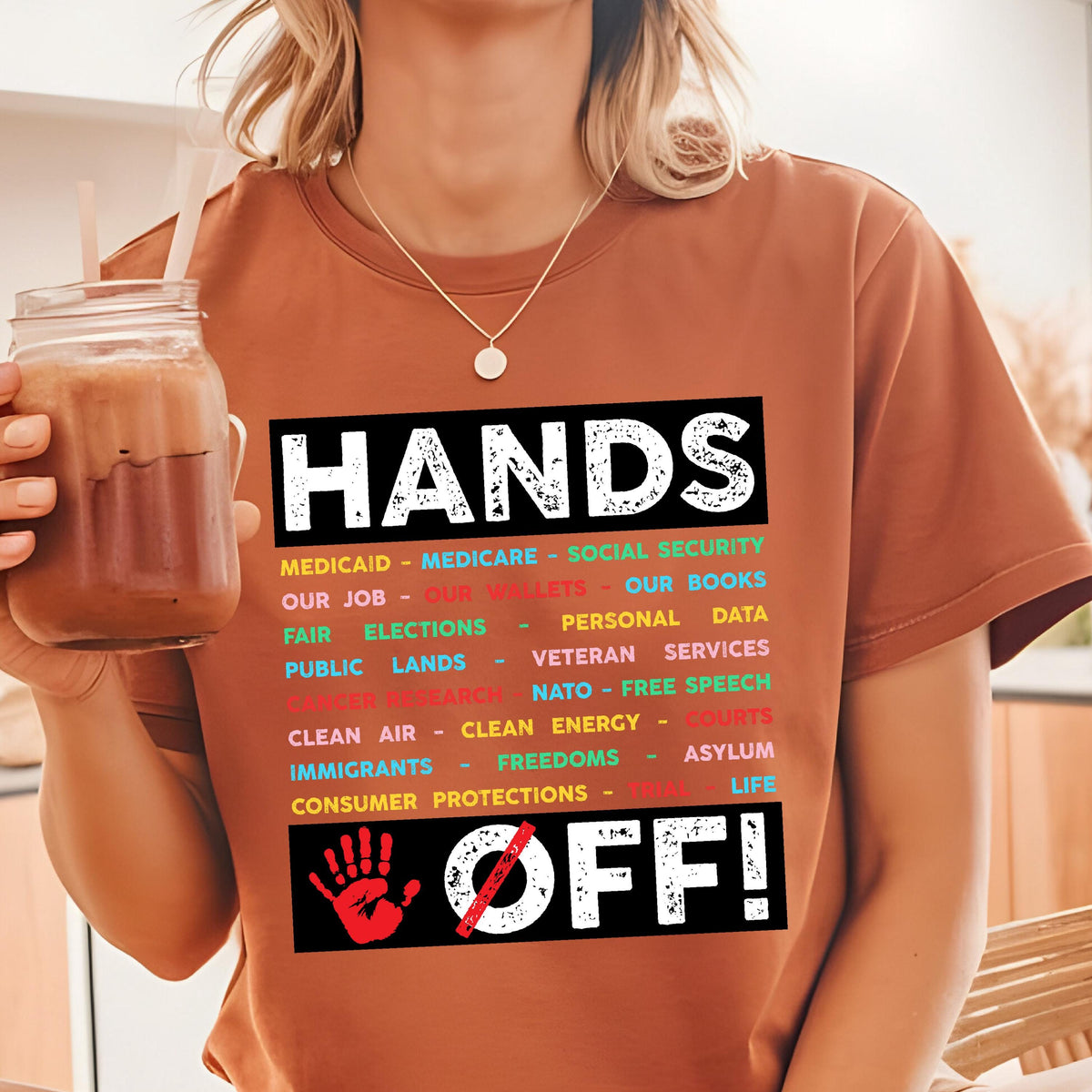 May include: A brown t-shirt with the text "HANDS OFF!" in black and white. The text is surrounded by a list of items in colorful text, including "Medicaid", "Medicare", "Social Security", "Our Job", "Our Wallets", "Our Books", "Fair Elections", "Personal Data", "Public Lands", "Veteran Services", "Clean Air", "Clean Energy", "Courts", "Immigrants", "Freedoms", "Asylum", and "Consumer Protections". A red handprint with a diagonal line through it is below the text.