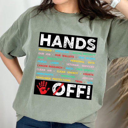 May include: A green t-shirt with the text "HANDS OFF!" in black and white. The text is surrounded by a list of words in different colors, including "Medicaid", "Our Job", "Our Wallets", "Personal Data", "Public Lands", "Veteran Services", "Cancer Research", "NATO", "Clean Air", "Clean Energy", "Courts", "Immigrants", "Asylum", "Consumer Protections", and "Trial". A red handprint with a diagonal red line through it is below the text.