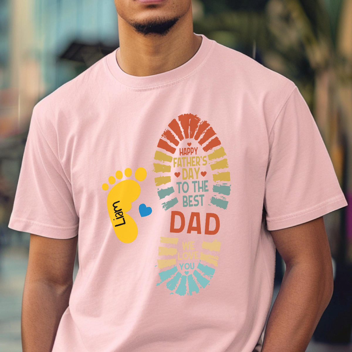 May include: Pink t-shirt with a colorful graphic for Father's Day. The design includes a footprint, a baby footprint, a blue heart, and the text "Happy Father's Day, To The Best Dad, We Love You". The baby footprint has the name "Liam".