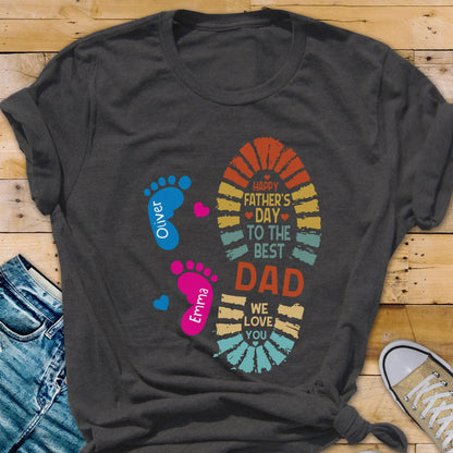 May include: Dark gray t-shirt with a colorful graphic for Father's Day. The design includes a large footprint with the text "Happy Father's Day to the Best Dad" and two smaller footprints with names. The shirt is made of soft material.