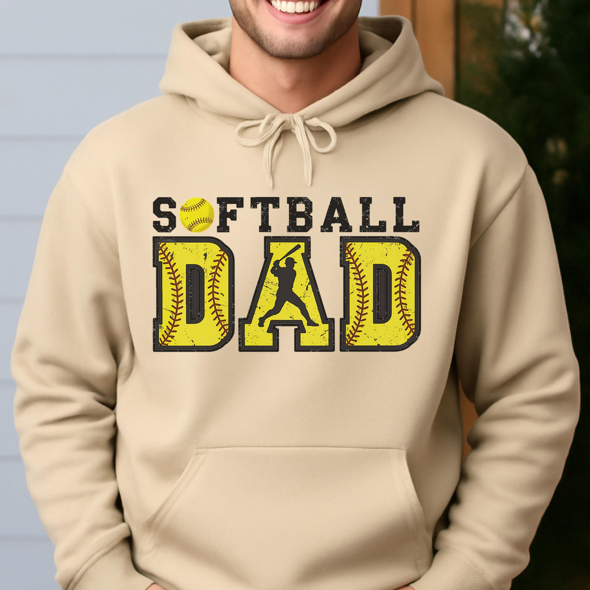 May include: A tan hoodie with the words "SOFTBALL DAD" in yellow and black. The word "DAD" features a silhouette of a batter. A yellow softball is above the word "SOFTBALL".