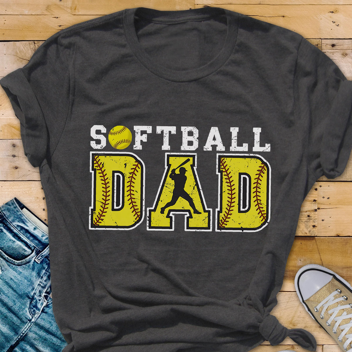 May include: A dark gray t-shirt with the text "Softball Dad" in white letters. The text is surrounded by yellow baseballs with red stitching. A silhouette of a softball player is in the middle of the text.