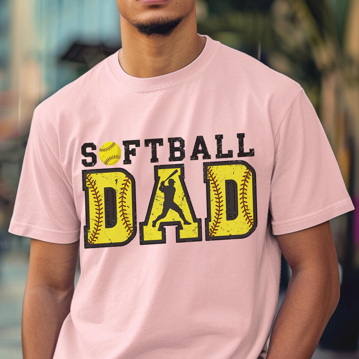 May include: Pink t-shirt with the text "Softball Dad" in black letters. The text is surrounded by two yellow softball graphics with red stitching. A silhouette of a softball player is in the middle of the text.