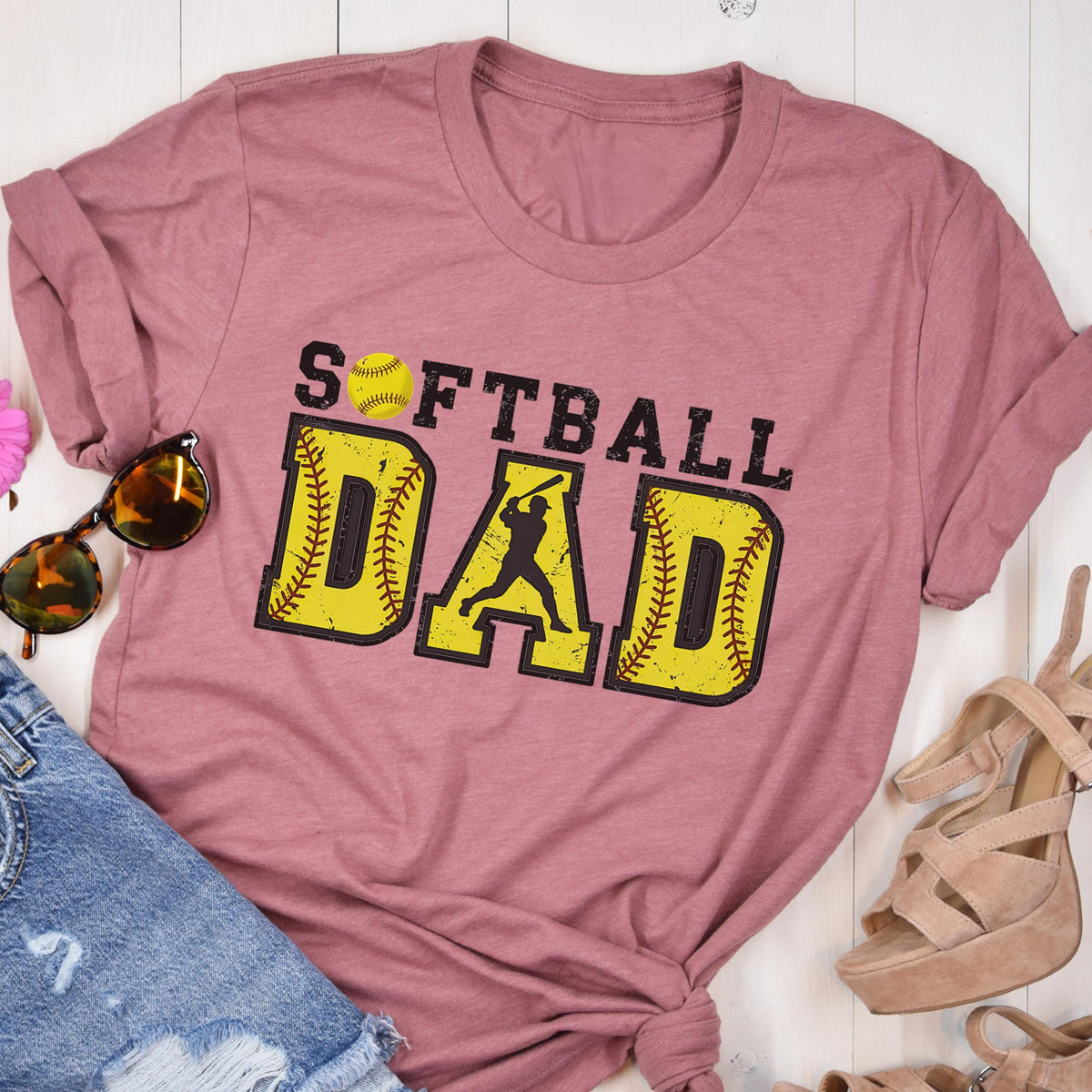 May include: A pink t-shirt with the text "Softball Dad" in yellow and black letters. The text is surrounded by two yellow baseballs with red stitching. A silhouette of a softball player is in the middle of the word "Dad".